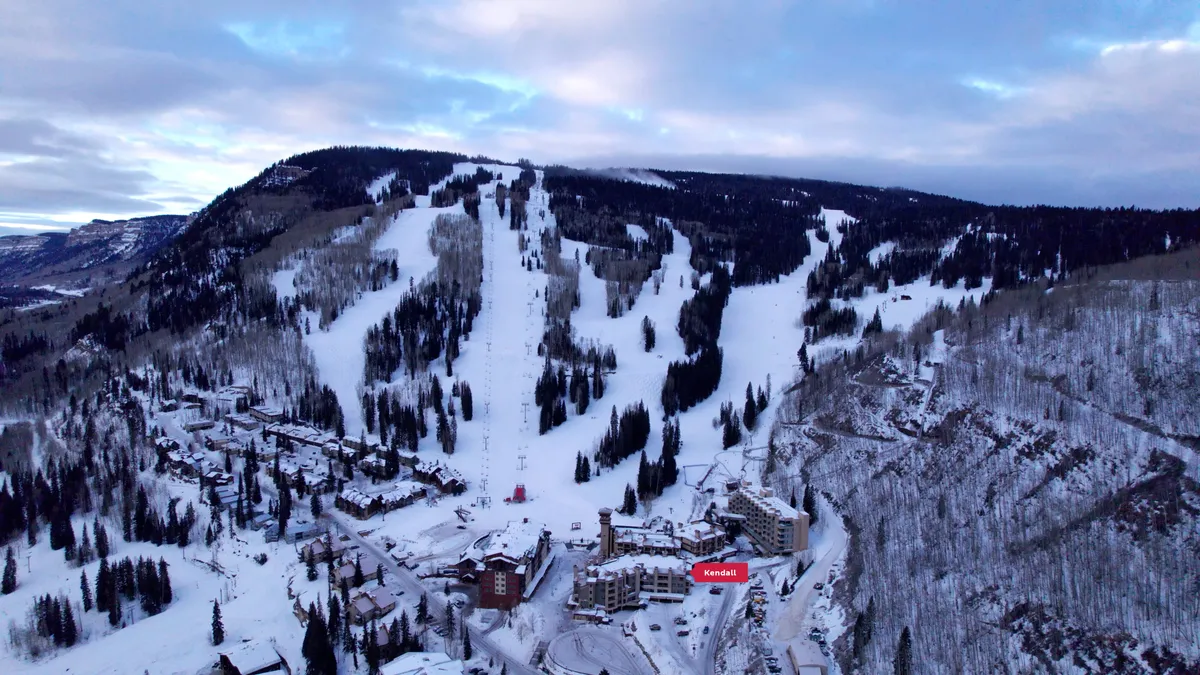 A panoramic view showcases the ski slopes, snow-dusted mountains, and cozy resort buildings, all drenched in invigorating natural light. The vantage point emphasizes both adventure and relaxation, connecting visitors to the dramatic landscape and the heart of resort life.