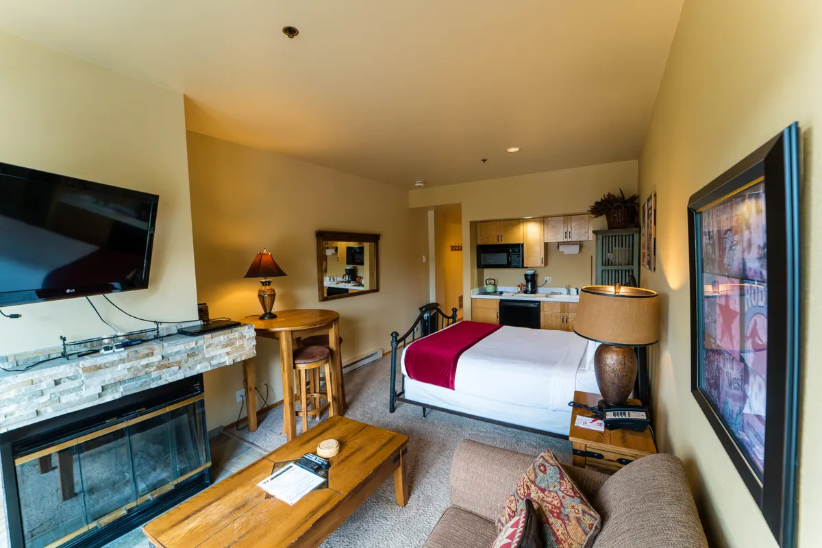 The image shows a studio living space featuring a Murphy bed placed near a glowing fireplace, a cozy lamp, and a compact kitchenette. The arrangement emphasizes warmth and multifunctionality, with the bed and seating area seamlessly blending into one cozy space.