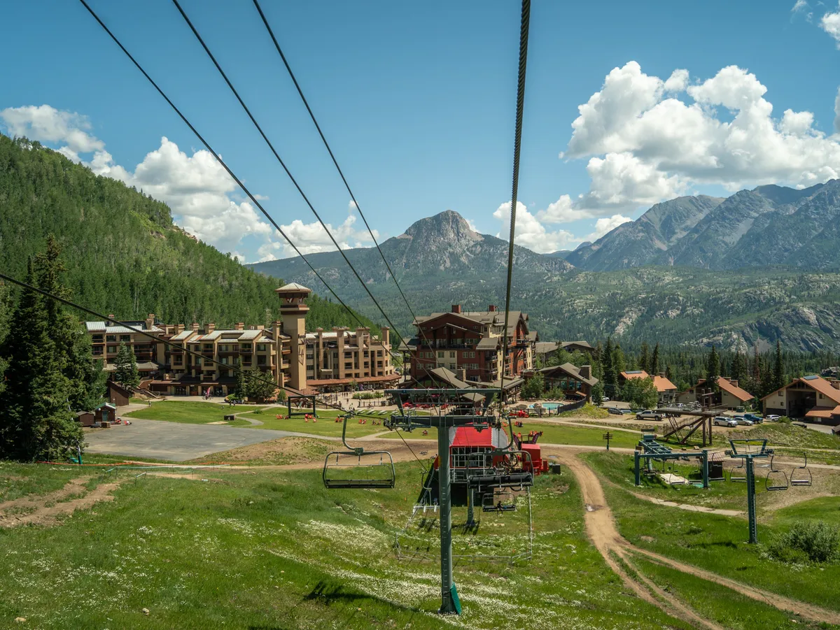 This exterior image features the ski resort buildings framed by a striking mountain backdrop, with lush greenery in the foreground. A ski lift is visible, highlighting access to outdoor adventures, while the tranquil setting evokes the resort's peaceful natural surroundings.