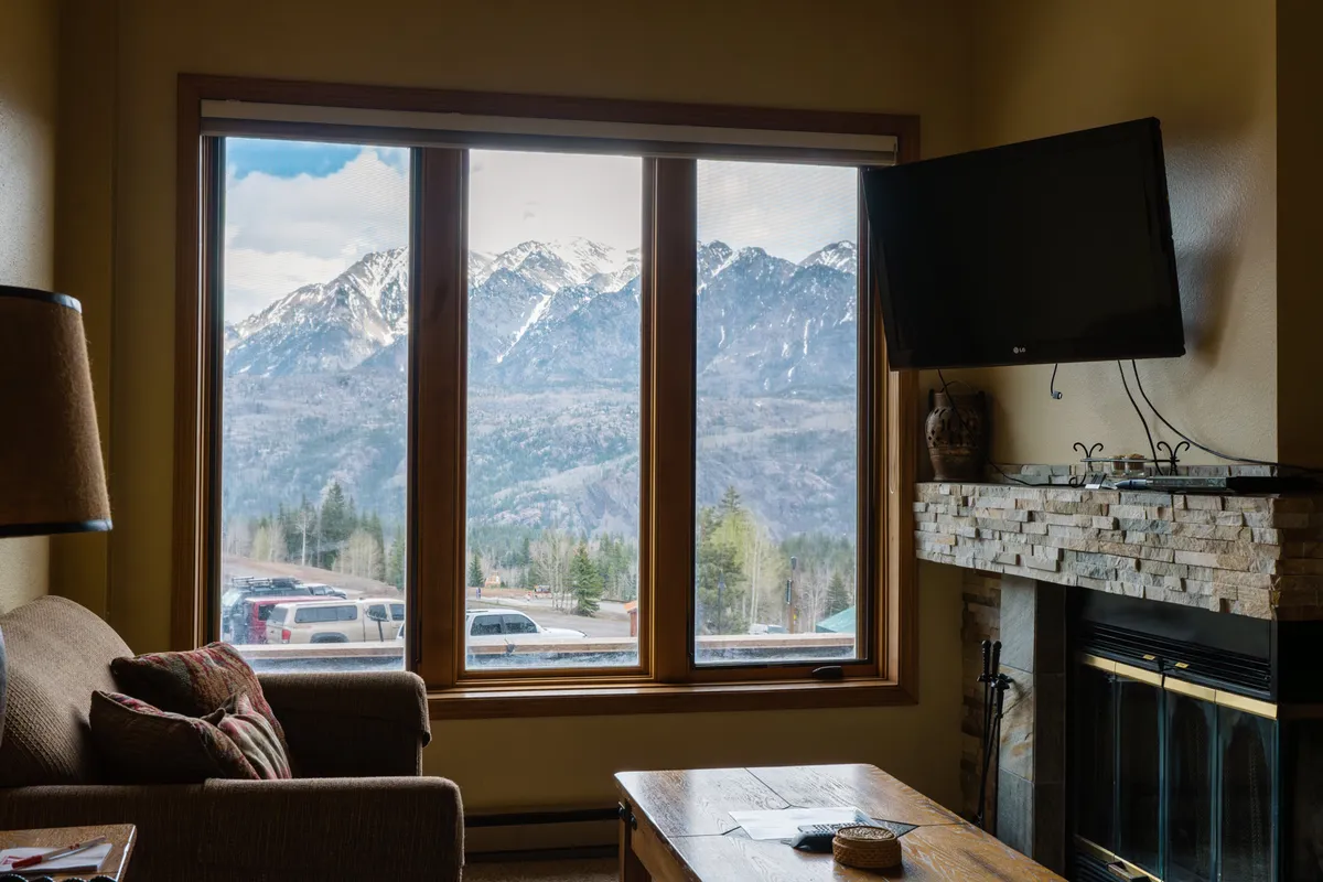 The image showcases a cozy condo interior with a comfortable couch facing a warm, inviting fireplace. Large windows frame breathtaking mountain views, flooding the room with natural light and offering the perfect spot to relax and watch the sunset over the Rockies.